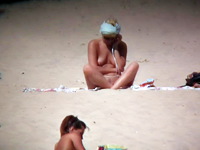 The nudist woman is sitting on the beach and talking on the phone. She does not notice somebody with cam is shooting her on camera